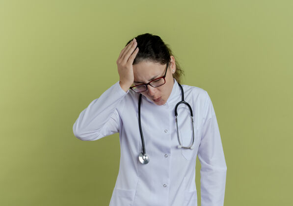 穿年轻的女医生戴着医用长袍和听诊器 戴着眼镜 手放在头上 眼睛紧闭 被隔离在橄榄绿的墙上放医学年轻