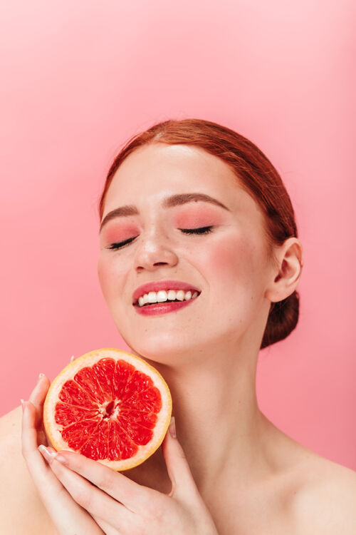 女性迷人的欧洲女孩拿着多汁的葡萄柚摄影棚拍摄了一个快乐的笑着的女人在粉红色背景下闭着眼睛摆姿势年轻成人葡萄柚表情