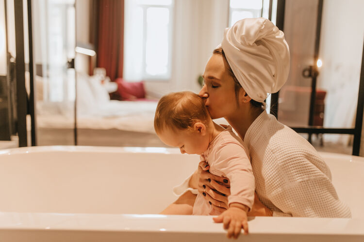 女儿穿着白色家装的漂亮女士亲吻她的孩子妈妈和宝宝坐在浴室里公寓淋浴浴缸