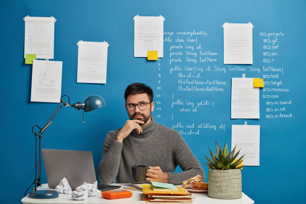 坐着穿着休闲套头衫 满脸胡须 沉思着公司信息 端着茶杯 在同事的空间里摆姿势 坐在蓝色背景下的笔记本电脑前桌面离开思考