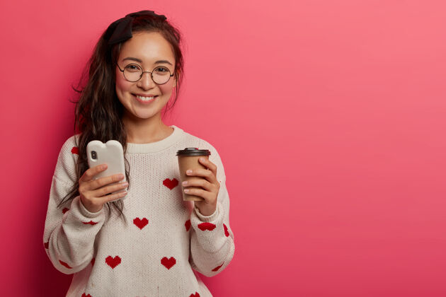 设备科技 通讯和生活理念戴着圆眼镜的漂亮女孩用智能手机阅读酷博客和信息 喝外卖咖啡 下载应用程序 站在室内亚洲人浏览马尾辫