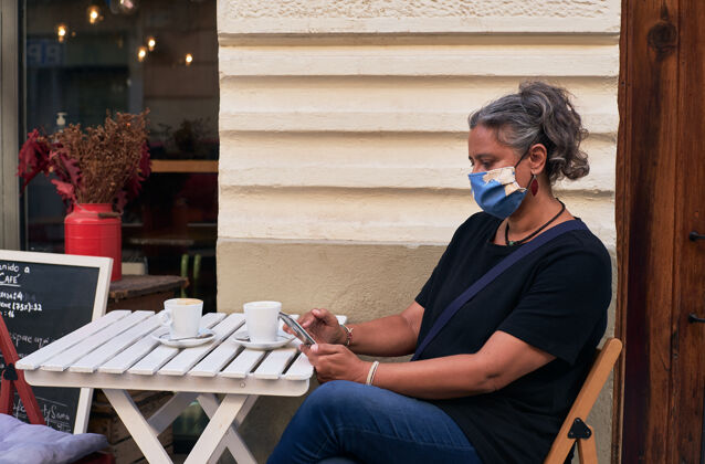 手一个戴着口罩的女人在咖啡馆的户外桌子上使用手机的侧视图桌子科技新闻