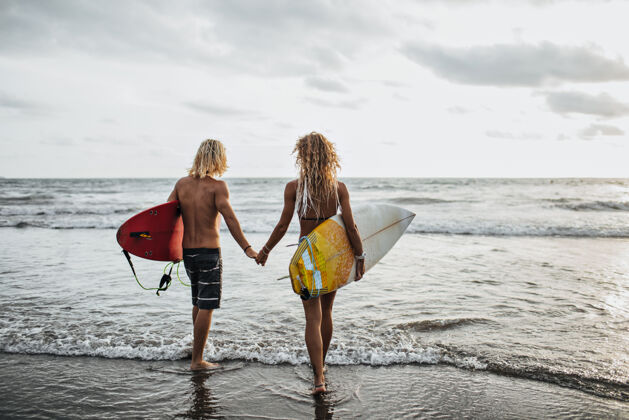 男性有着波浪形头发的男孩和女孩手牵着冲浪板去海边夏天感情浪漫