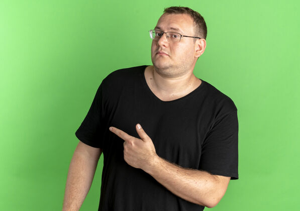 男士戴眼镜的超重男子身穿黑色t恤 看上去很困惑 食指指向绿色上方的一侧困惑站壁板