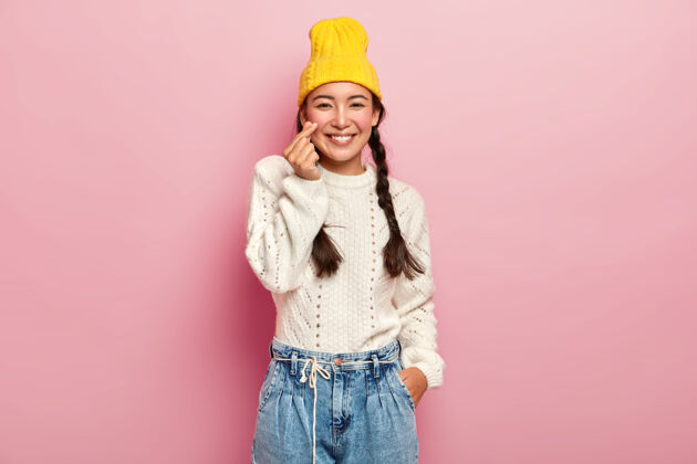 牛仔裤漂亮的亚洲女孩把手指叠成心形符号 展示韩国爱情符号 戴着时髦的黄色帽子 白色毛衣和牛仔裤 深色头发梳成两条辫子 对着粉色的墙壁摆姿势手势服装女