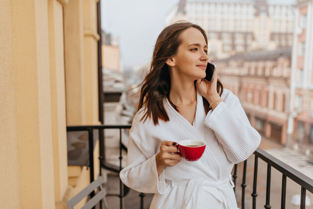 脸一位留着长发的年轻女子在阳台上欣赏着城市的景色穿着浴衣的女孩一边喝咖啡一边打电话黑发女性室内