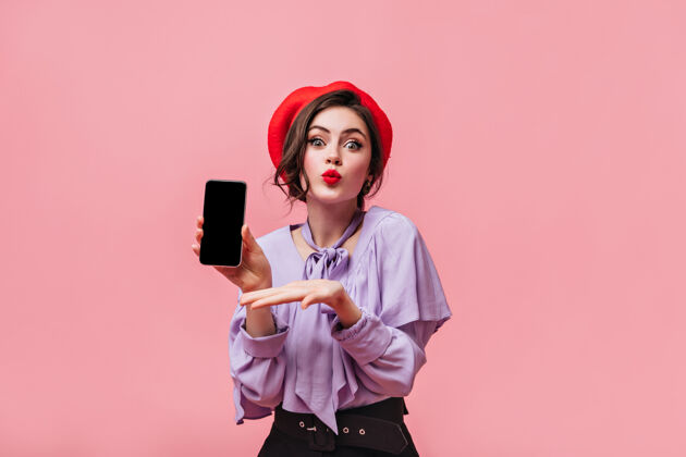 肖像绿色眼睛的年轻女孩在粉色背景上演示智能手机戴着红帽子和带褶边的淡紫色衬衫的女士肖像裤子黑色年轻