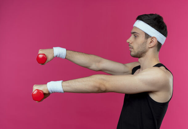 站立年轻的运动型男子戴着头巾 戴着哑铃 紧张而自信地在粉色的头上锻炼工作自信头带