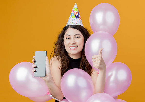 节日年轻漂亮的女士 卷发 戴着节日帽 手持气球和智能手机 竖起大拇指 快乐而兴奋的生日派对理念超越橙色移动显示气球