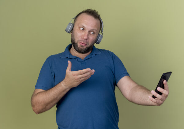 耳机印象深刻的成年斯拉夫男子戴着耳机拿着手机看着并用手指着男士指点手持