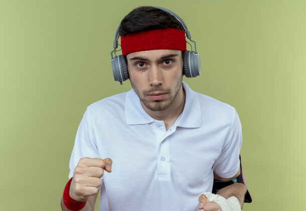 脸戴着头戴式耳机和智能手机臂带的年轻运动型男子握紧拳头 严肃的脸盖过绿色手臂拳头握紧