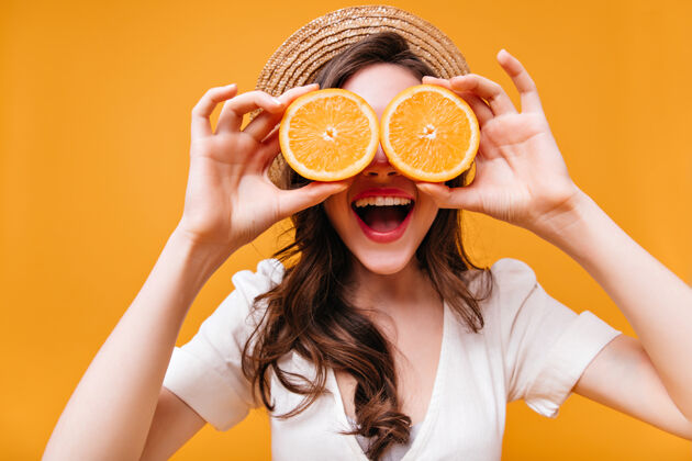 肖像穿着白色上衣戴草帽的女士笑着用橘子遮住眼睛波浪发黑发橙色背景
