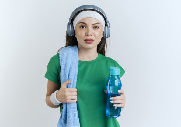 看自信的年轻运动女性戴着头带 腕带和耳机 肩上拿着毛巾拿着水瓶抓着毛巾看抓肩膀运动