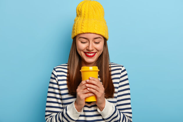 化妆品快乐的女人戴着时髦的黄色帽子和条纹套头衫 拿着外卖咖啡 涂着红色的嘴唇 享受着芳香的饮料自由时间站立外卖