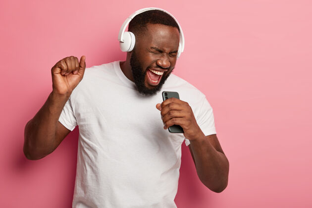 男孩精力充沛的黑胡子男人随着音乐唱歌 动作积极 戴着耳机和休闲t恤 在粉色背景下摆姿势 张大嘴巴欢呼大声表情