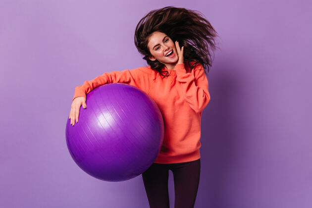 年轻人身着鲜艳运动衫和深色紧身裤的微笑女士手拿健身球跳上紫色墙壁女人锻炼身体