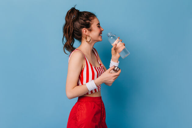时尚穿红条纹衣服的年轻女子笑着喝着隔着墙的瓶子里的水长美女红色裤子