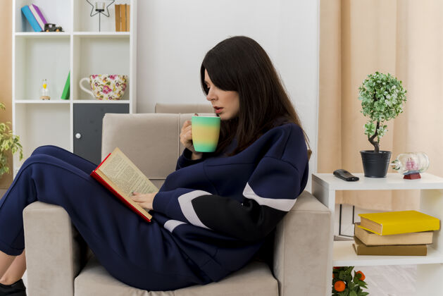 女人年轻漂亮的白人妇女坐在设计客厅的扶手椅上 手里拿着杯子 腿上放着书 抚摸着书 准备喝咖啡阅读房子腿