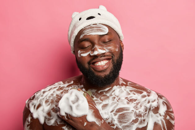 人放松的时间和淋浴的概念高兴的非洲男人愉快地闭上眼睛 笑容灿烂 在浴室用海绵清洗 应用泡沫凝胶护理年轻洗脸