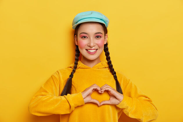 微笑年轻快乐的韩国女人在胸前做心形 留着两条辫子 戴着蓝色帽子和黄色连帽衫 表达美好的情感肖像穿孔女性