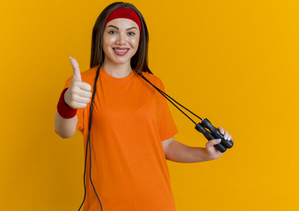 年轻微笑的年轻运动女性戴着头带和腕带 脖子上系着跳绳 抓着跳绳 向上伸出大拇指看向上运动