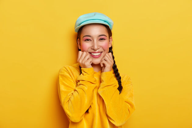 化妆令人愉快的亚洲女孩 双手放在下巴下 化着小妆 戴着蓝色时髦的帽子 丝绒黄色的连帽衫女性黑发高兴