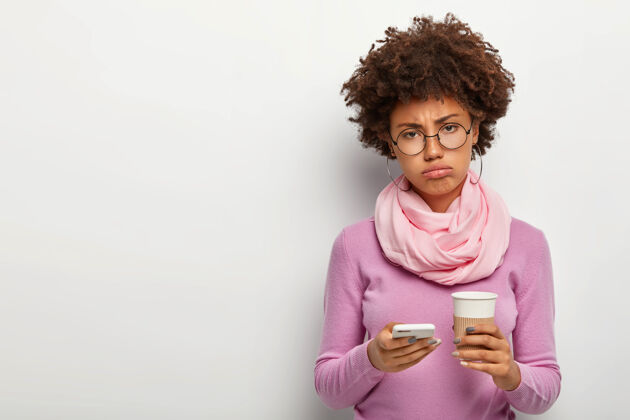 不满不高兴的悲伤的非洲女人厌倦了在办公室里的辛苦工作 拿着手机 下载新的应用程序 拿着外卖咖啡 戴着圆圆的大眼镜和围巾高领毛衣杯子打字科技