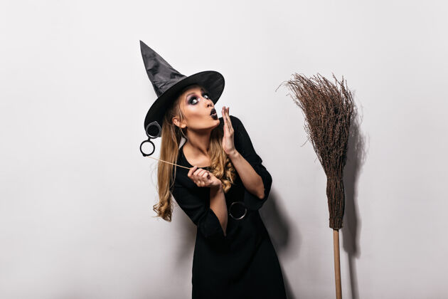 魔术在嘉年华里 一个穿着女巫服装的有趣的白人女孩戴着魔法帽的长发女人拿着扫帚站在白色的墙上神秘邪恶传统