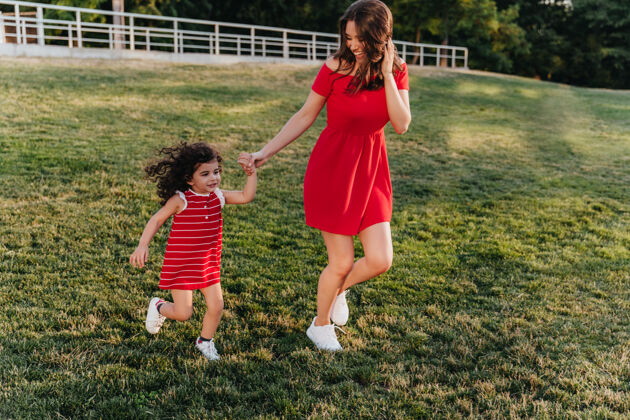 拥抱无忧无虑的年轻母亲牵着孩子在公园里跑来跑去穿着红裙子的滑稽女人和她的女儿在草地上跳舞表情草地放松