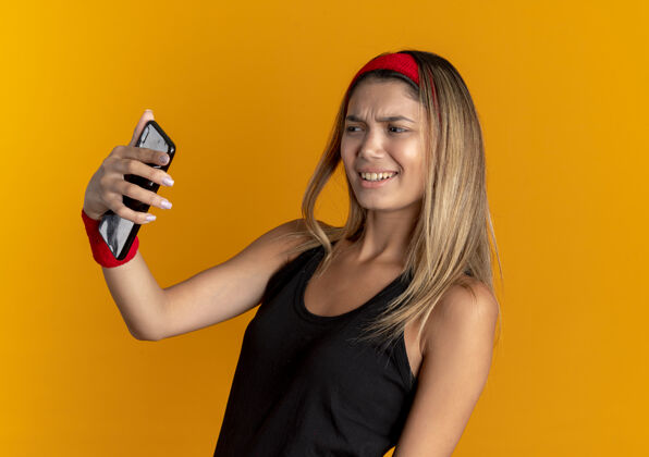 自拍身穿黑色运动服 头戴红色头巾的年轻健身女孩站在橘色的墙上用智能手机自拍 看起来很不高兴采取年轻头带