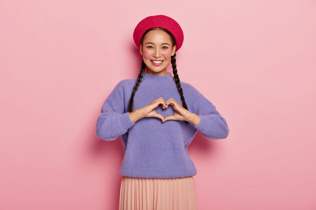 心外表迷人的年轻女士 在胸前做一个心形手势 承认爱情 戴着红色贝雷帽 紫色超大套头衫 化着妆 隔离在粉红色的墙上微笑套头衫休闲