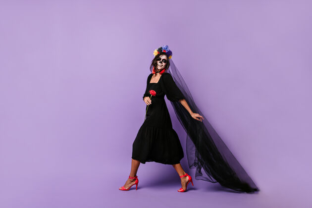 新娘身着万圣节服装的墨西哥笑脸模特手持长长的黑色面纱 穿过淡紫色的墙壁僵尸颜料人物