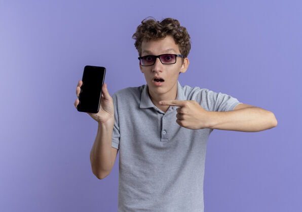 马球一个戴着黑眼镜 穿着灰色马球衫的年轻人拿着智能手机 用食指指着站在蓝色墙壁上的那个人手指拿着惊喜
