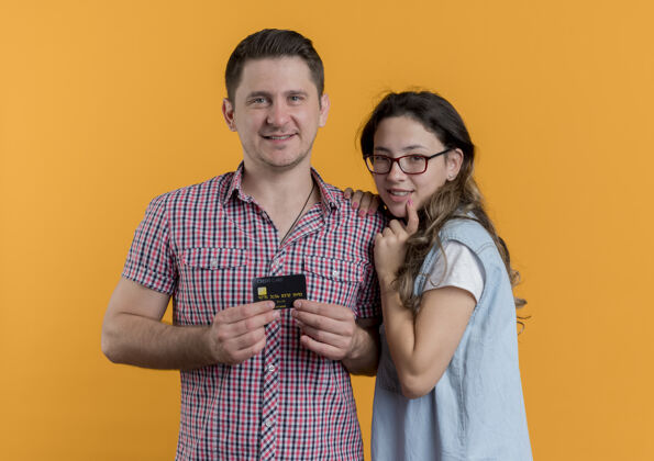 衣服一对穿着休闲服的年轻夫妇快乐的男人拿着信用卡站在他女朋友旁边橙色的墙上显示女人信用