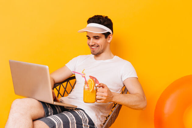 冒险一个穿着白色t恤的男人度假时在笔记本电脑里工作的肖像这个家伙喝的是橙色鸡尾酒椅子太阳镜行李