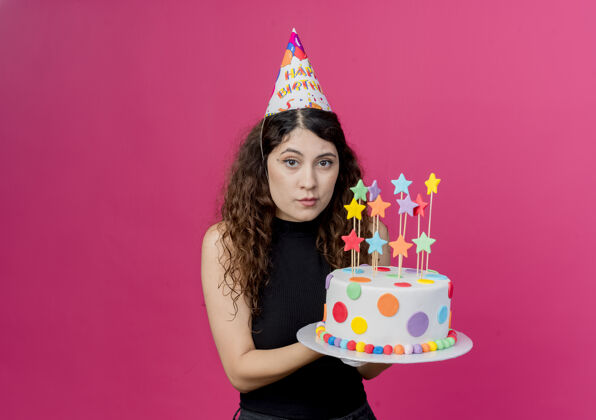 卷发年轻漂亮的女人 卷发 戴着节日帽 手里拿着生日蛋糕 严肃的脸 站在粉色的墙上 生日派对的概念抱着帽子脸