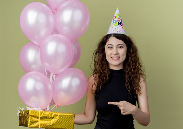 气球一位年轻漂亮的女士 头上戴着节日帽 手里拿着气球和礼盒 手指着礼盒 微笑着 站在明亮的墙上 快乐而积极女人积极微笑