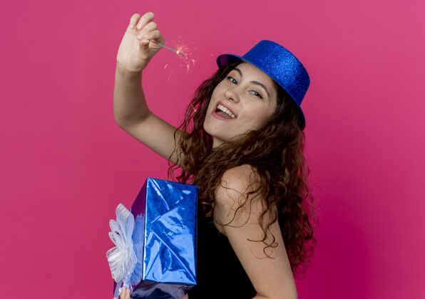 兴奋一位年轻漂亮的女士 卷发 戴着节日礼帽 手拿生日礼盒 手里拿着闪闪发光的生日礼物 站在粉色的墙上 快乐而兴奋的生日派对概念闪闪发光年轻女人