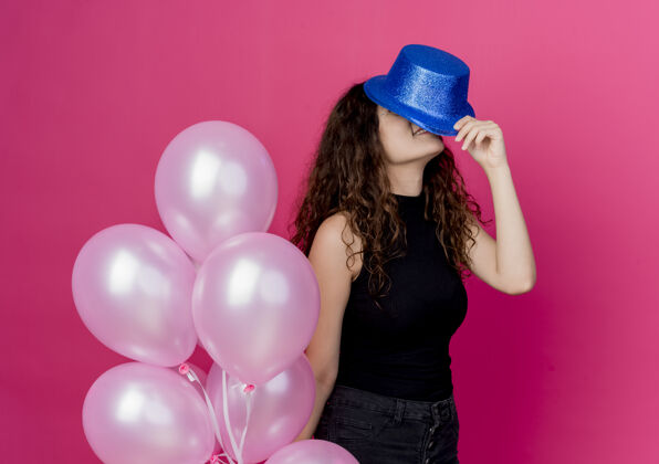帽子一个年轻漂亮的女人 一头卷发 戴着节日帽 手里拿着一堆气球站在粉红色的墙上年轻女人气球