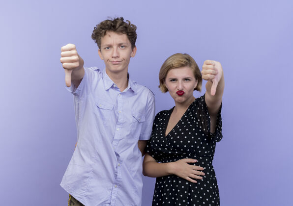 显示一对年轻漂亮的夫妇不高兴地在蓝色的墙上竖起大拇指男人女人拇指