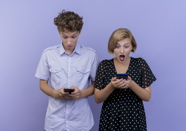 情侣一对年轻漂亮的夫妇站在蓝色的墙上惊讶地看着他们的智能手机惊喜移动站