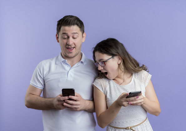 小玩意一对幸福的年轻夫妇 男人和女人在使用电子产品 女人低头看着她男朋友的智能手机屏幕 站在蓝色的墙上使用年轻情侣