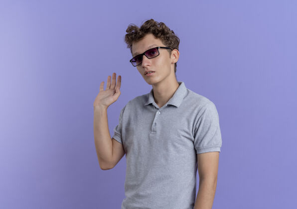 年轻一个戴着黑眼镜 穿着灰色马球衫的年轻人站在蓝色的墙上挥手致意站手看