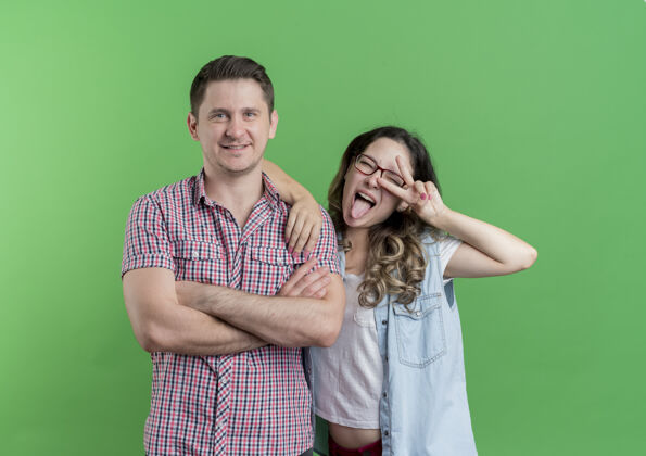 女人一对穿着休闲服的年轻男女快乐的男人站在他微笑的女友旁边 在绿色的墙上展示着v形标志女朋友Vsign男人