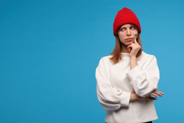 情绪严肃的 深色头发的成年女孩的肖像穿着白色毛衣和红色帽子用手指触摸她的脸颊和思考在复制空间的左边看 隔离在蓝色的墙上外观脸颊思考