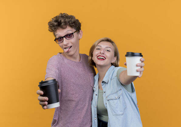 微笑一对年轻漂亮的情侣穿着休闲服展示咖啡杯微笑着站在橙色的墙上男人穿着女人