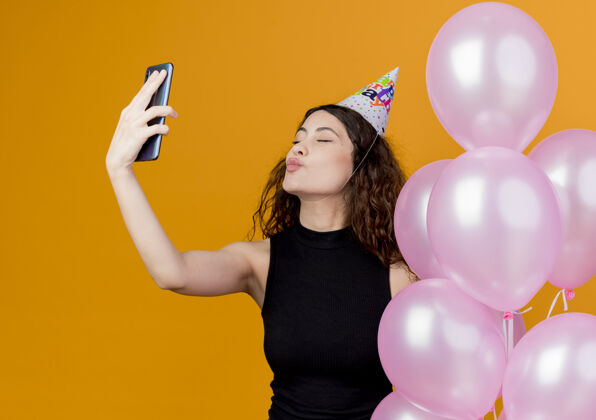 自拍一个年轻漂亮的女人 卷发 戴着节日帽 手里拿着气球吹着吻 自拍生日派对 站在橙色的墙上拿着站着空气