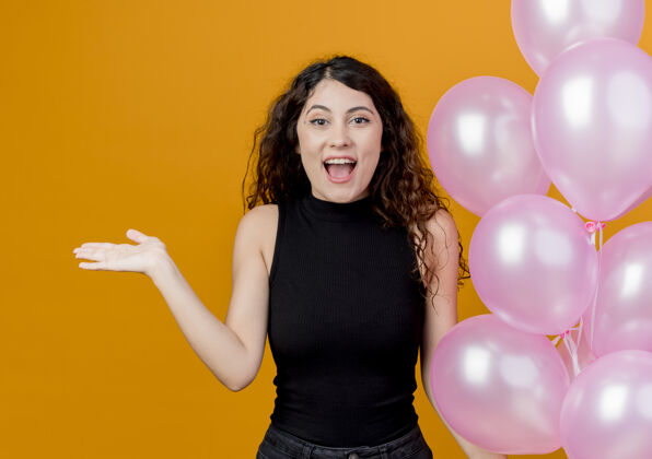 女人年轻漂亮的卷发女子手持一束气球站在橙色的墙上 快乐而兴奋的生日派对概念看着空气站着
