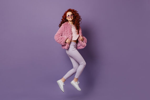 太阳镜穿着时尚紧身裤 白色t恤和粉色外套的迷人女孩在淡紫色的空间跳跃奢华淑女波浪发
