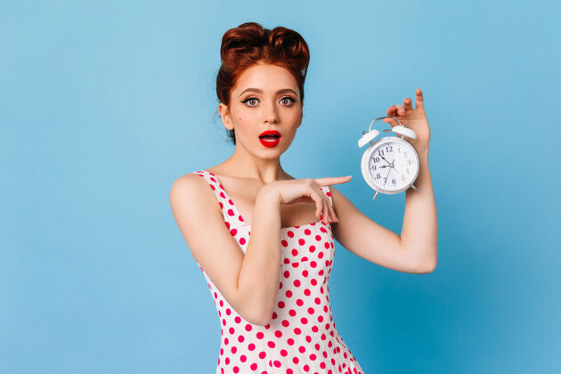 搞笑惊艳的女人带着明亮的妆容展示时间摄影棚拍摄的蓝色空间里拿着时钟的惊艳的小女孩物体欧式时钟
